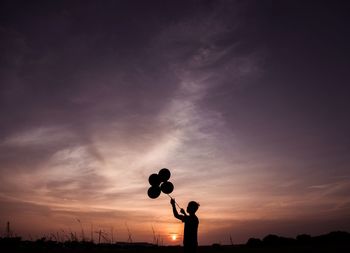 Silhouette boy holding balloons against sky during sunset