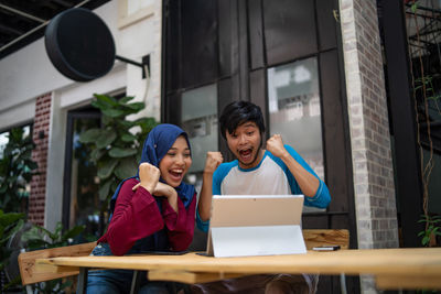 Low angle view of cheerful couple using digital tablet while sitting at sidewalk cafe