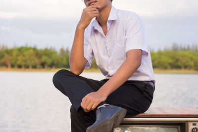 Teenage boy sitting on television set at lakeshore against sky