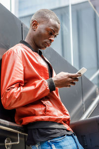 Side view of concentrated young african american guy in trendy red leather jacket messaging on mobile phone while leaning on wall on city street