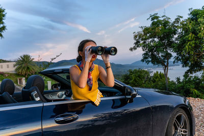 Young woman in yellow dress looking through binoculars from car