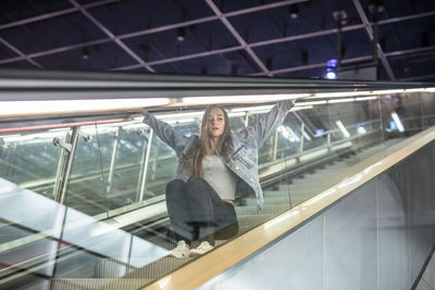 Young woman looking away while sitting on escalator seen through glass