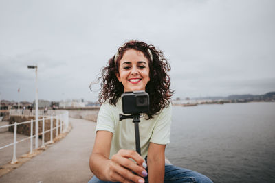 Smiling woman blogging while sitting by sea against sky