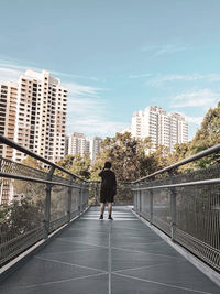 Rear view of man walking on footbridge in city against sky