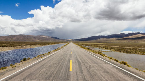 Country road passing through landscape against cloudy sky