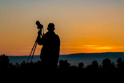 Silhouette of man with camera standing against sky during sunset