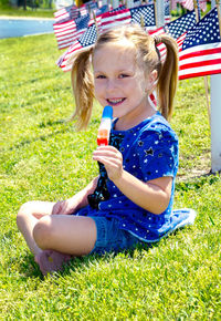  little girl eats a red white and blue popsicle, as she sits on a hill surrounded by american flags