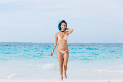 Full length of smiling woman in bikini standing at beach against sky