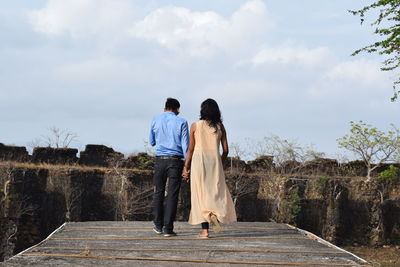 Rear view of couple holding hands while walking on boardwalk against sky