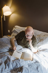 Senior man with hand on chin reading book while lying on bed at home