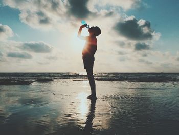 Side view of man standing and drinking water at sea shore against cloudy sky