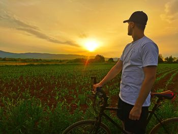 Man standing by bicycle on field against sky during sunset