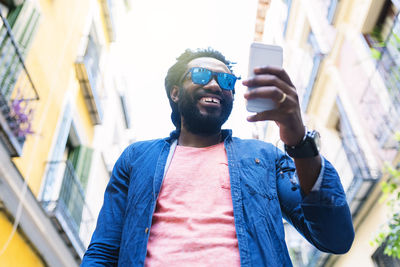 Low angle view of smiling fashionable man using smart phone in city