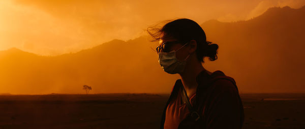 Woman wearing face mask standing against orange sky at sunset