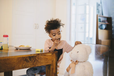 Boy eating food while looking at teddy bear in brightly lit room