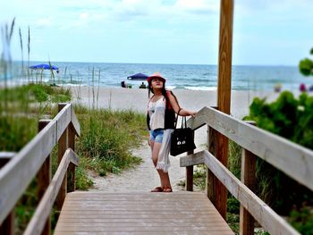 Fashionable woman standing on footbridge at beach against sky