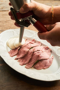 From above of faceless chef adding fluffy sauce on plate with slices of gentle roast beef meat on wooden table