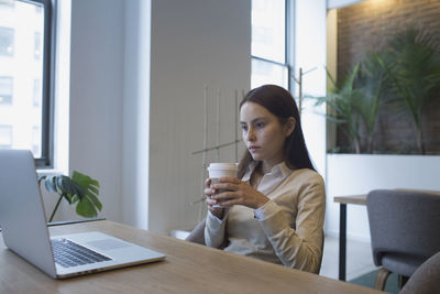 Young woman sitting at desk with coffee while looking at laptop computer.