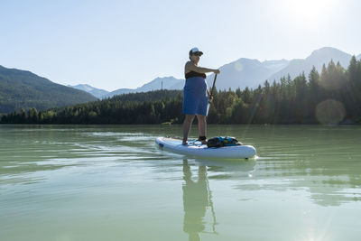 Full body pregnant woman in dress paddleboarding on calm water of lake against mountain ridge and green forest on summer day in british columbia, canada
