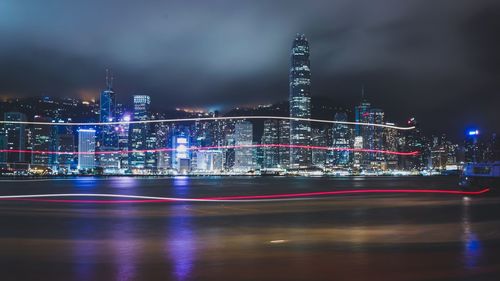Light trails over river against illuminated cityscape at night