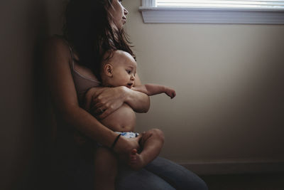 Mother looking through window while holding shirtless son by wall at home