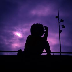 Silhouette boy sitting on railing against sky during sunset