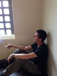 Side view of young man smoking cigarette while sitting against wall at home