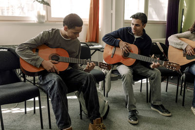 Teenage boys practicing guitar sitting on chair in music class at high school