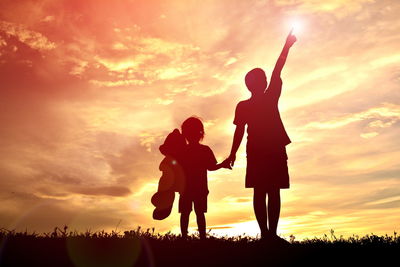 Silhouette girl holding teddy bear with brother on field against sky during sunset