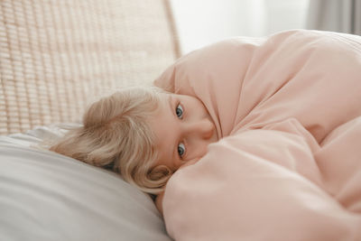 Pensive caucasian blonde child girl lying on pillow under blanket in bed at home. sad kid in stress 