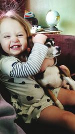 Portrait of cute smiling girl stroking dog while sitting on sofa at home