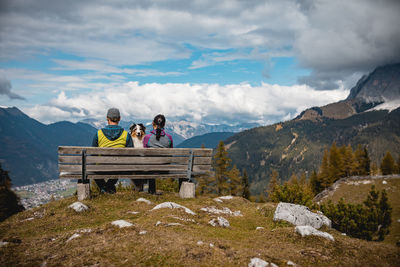 Couple with dog sitting on bench by mountains against cloudy sky