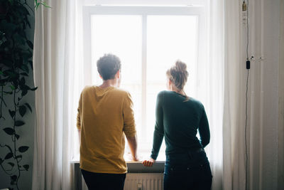 Rear view of friends looking through window at home