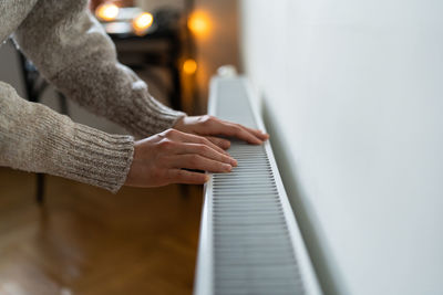 Closeup of woman in sweater puts hands on room central heating battery to warm up in cold apartment