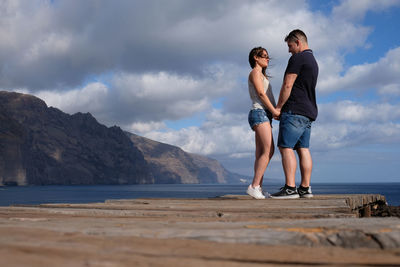 Full length of couple standing on pier over sea against cloudy sky