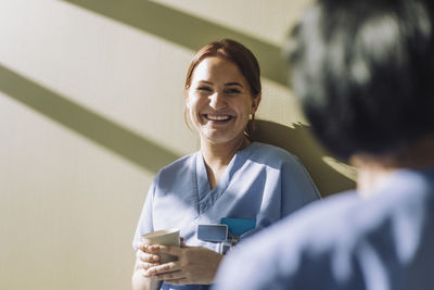 Smiling female medical trainee holding disposable cup near wall at hospital