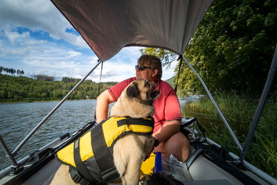 Pug dog with life jacket in a rubber dinghy. the owner sits with in the boat and looks at the lake.