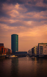 Sea by modern buildings against sky during sunset