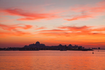 Silhouette buildings by sea against dramatic sky during sunset