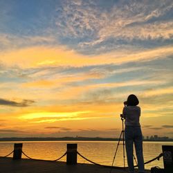 Rear view of woman photographing by sea against sky during sunset