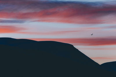 Silhouette bird flying over mountains against orange sky