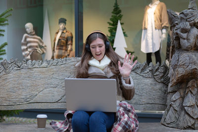 A young woman with headphones on and a laptop greets someone while sitting on a bench 