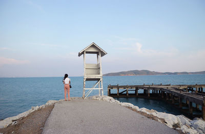 Rear view of woman standing by lifeguard hut and sea against sky