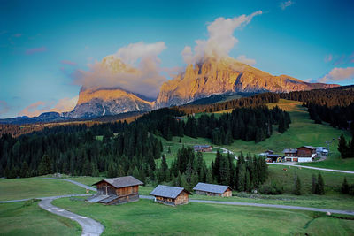 Panoramic view of alpine pasture and woods with huts against dolomite mountains at sunset. 