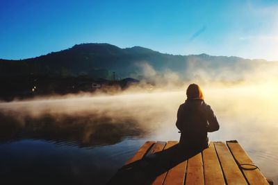 Rear view of woman sitting on jetty by lake against clear sky