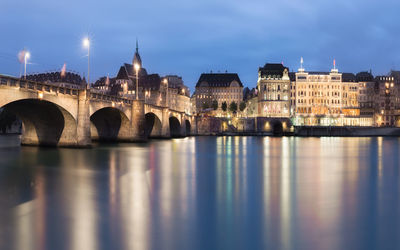 Bridge over river by illuminated buildings against sky at night