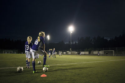 Girls practicing soccer on field against sky at night