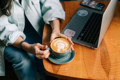 Female hands hold a large mug with cappuccino at a table in a cafe next to a laptop.