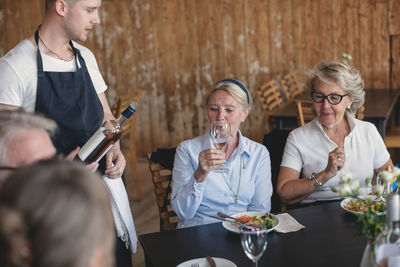 Waiter looking at senior woman drinking wine while having lunch with friends at restaurant