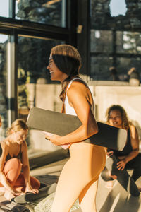 Side view of happy female coach holding exercise mat at retreat center